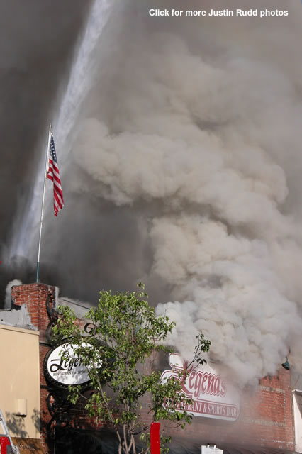 The American Flag stands tall as billows of smoke rise during the downpour as a fire hose douses the burning building.